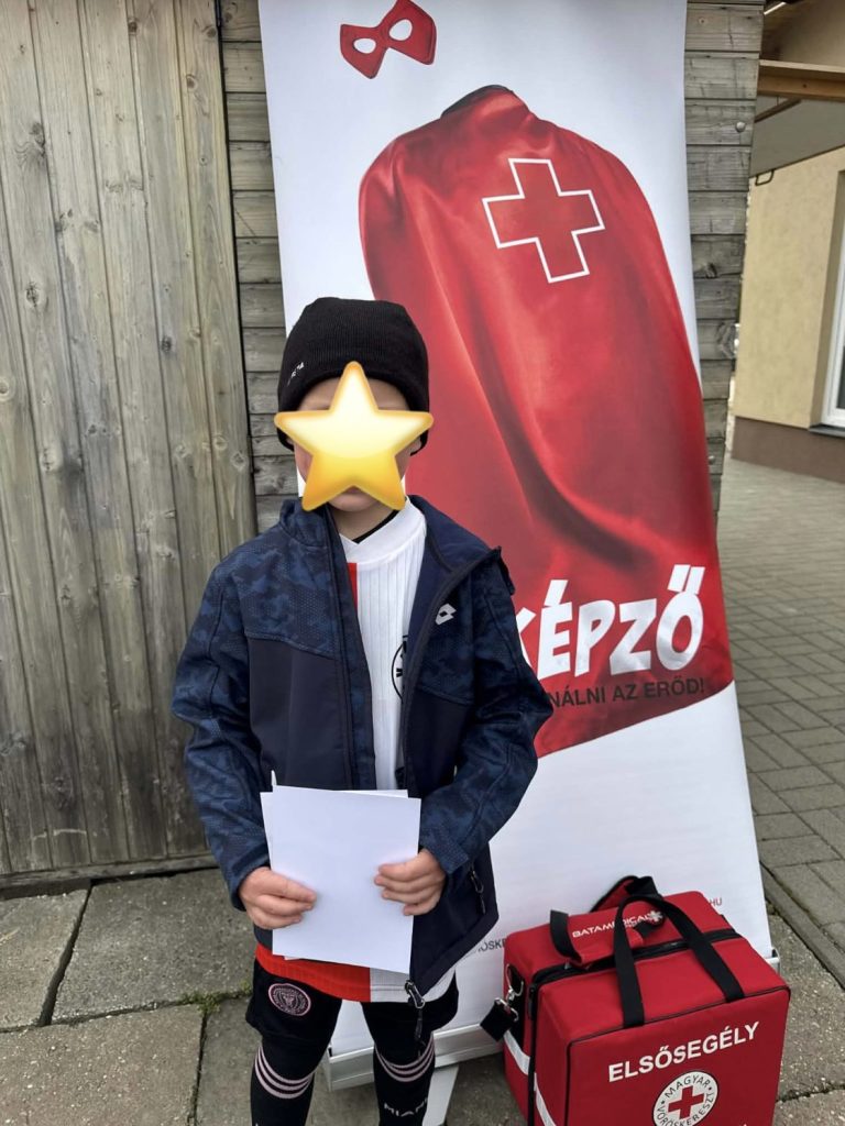 Gyermek áll a Magyar Vöröskereszt piros elsősegély-táskája mellett egy vöröskeresztes molinó előtt