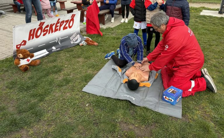 Vöröskeresztes önkéntes újraélesztést mutat be egy gyermeknek egy babán a Bozsik Fesztiválon, mellette a Hősképző molinó